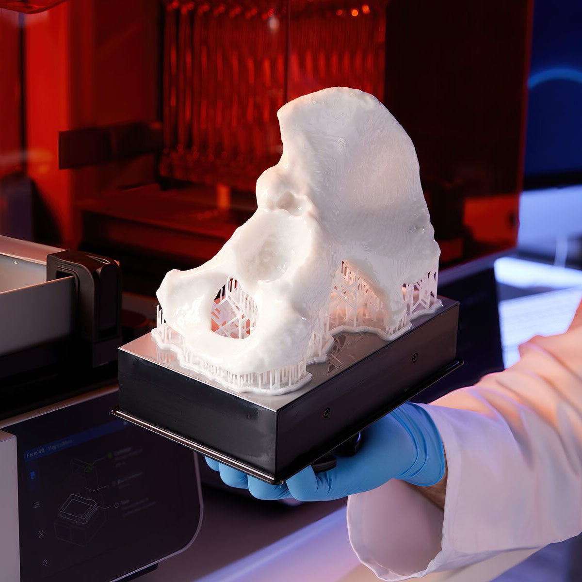 3D-printed object held by a person wearing blue gloves in front of a 3D printer.