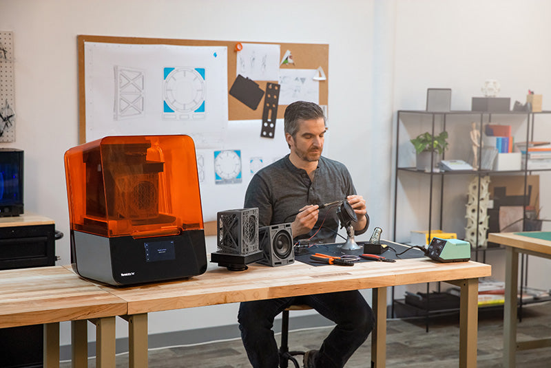 Man sitting in Additive Manufacturing workshop at a desk next to a 3D printer.